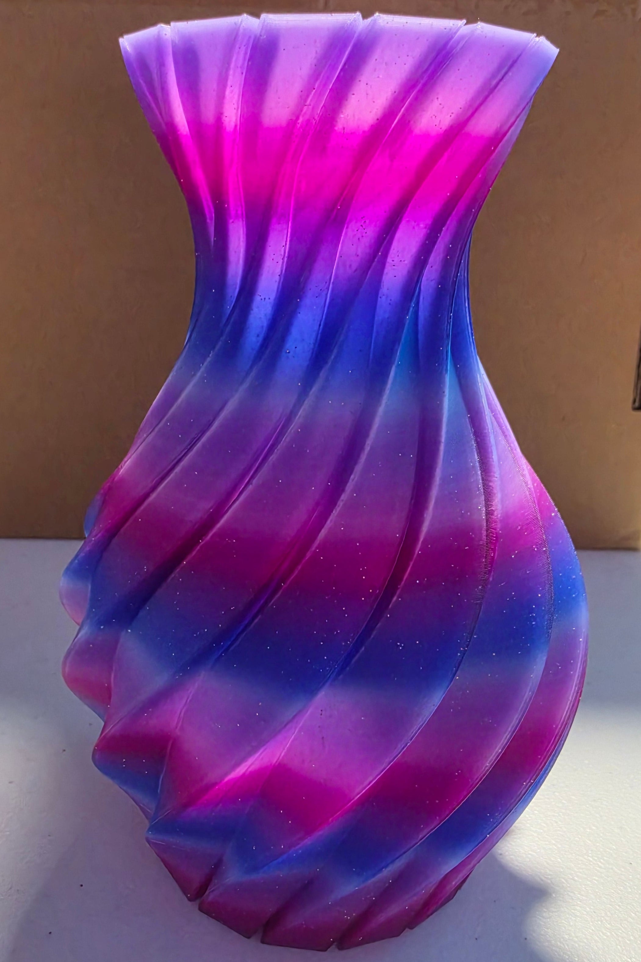 A twisted glass vase with a vibrant purple and blue gradient, standing in front of a brown cardboard box on a white surface.