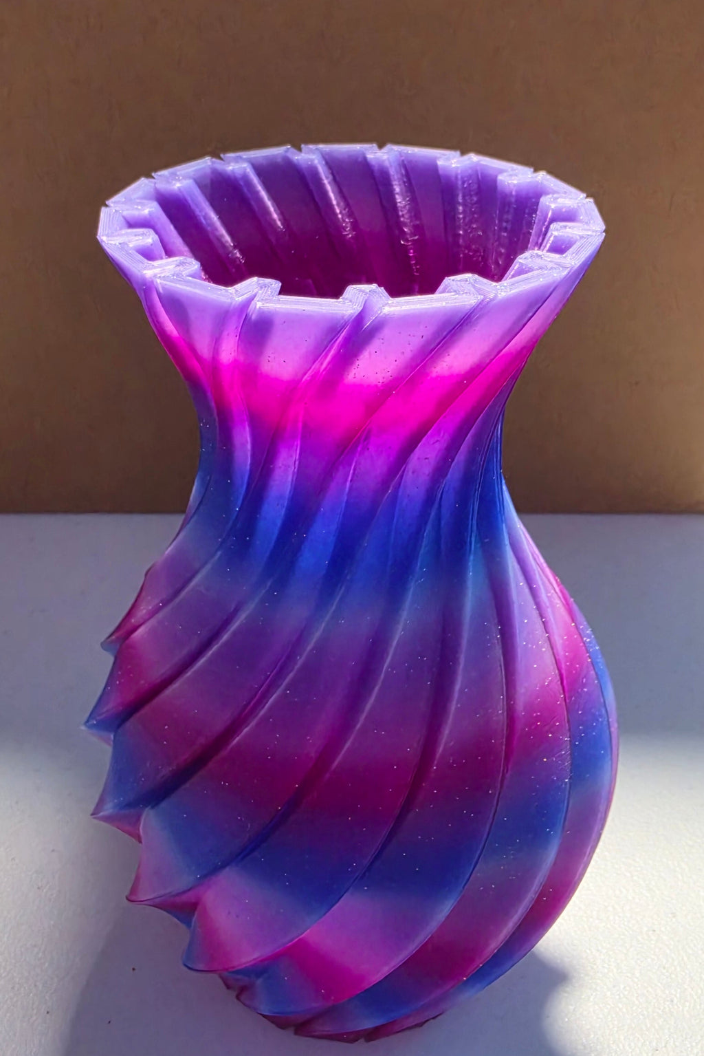 A twisted, flared vase with a vibrant purple and blue gradient, standing on a white surface in front of a brown cardboard box.