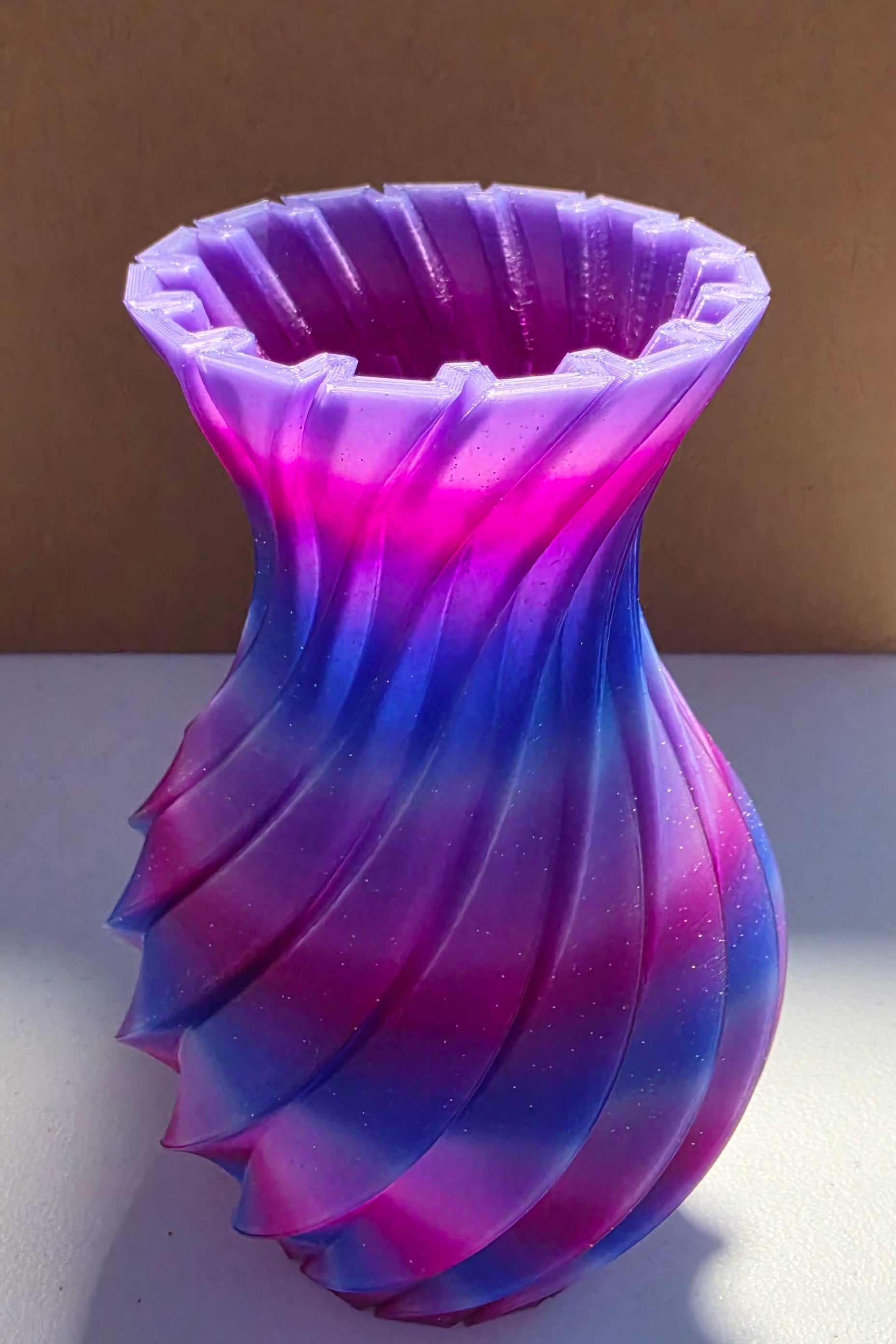 A twisted, flared vase with a vibrant purple and blue gradient, standing on a white surface in front of a brown cardboard box.