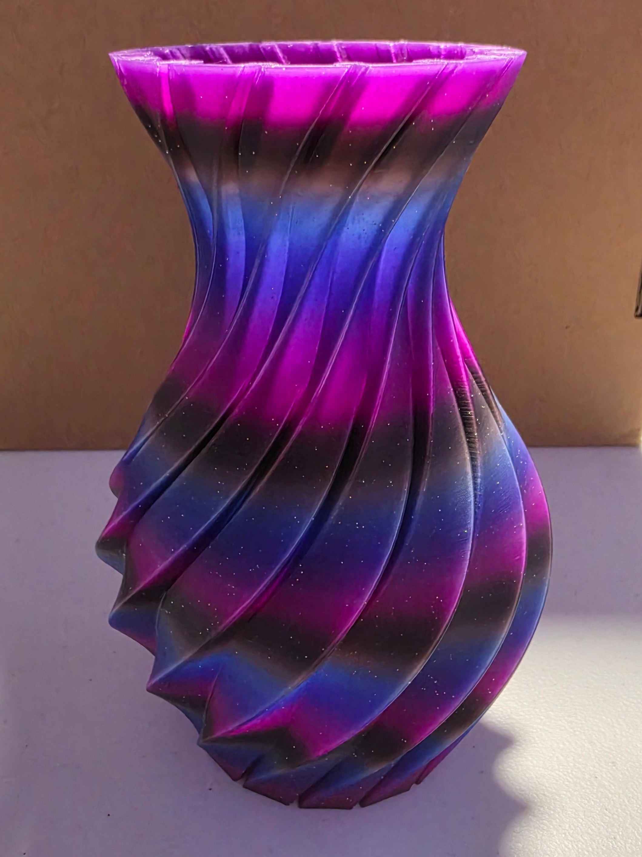 A 3D-printed twisted vase with a vibrant purple, blue, and black gradient pattern, standing on a white surface in front of a cardboard box.