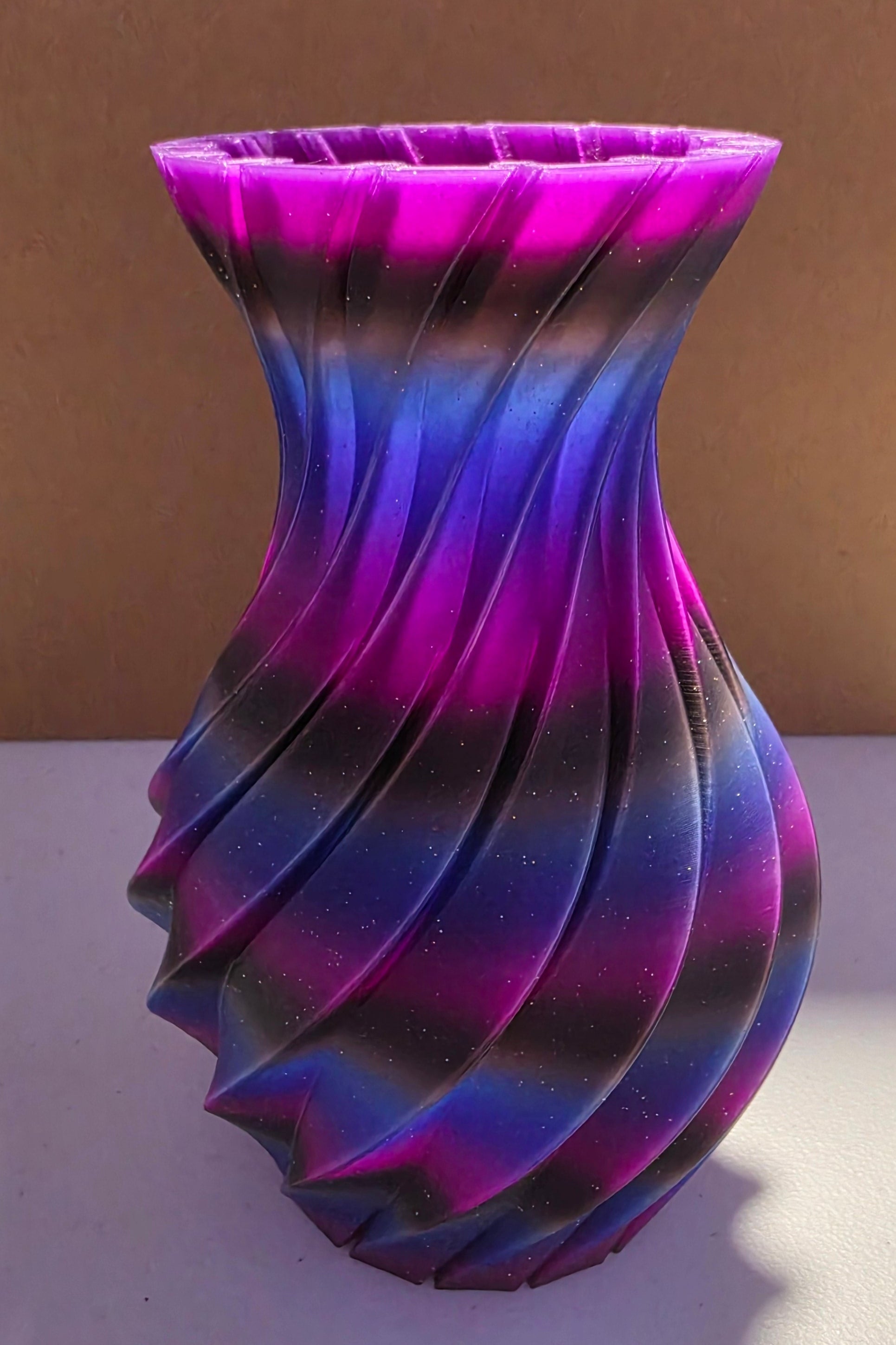 A 3D-printed twisted vase with a vibrant purple, blue, and black gradient pattern, standing on a white surface in front of a cardboard box.